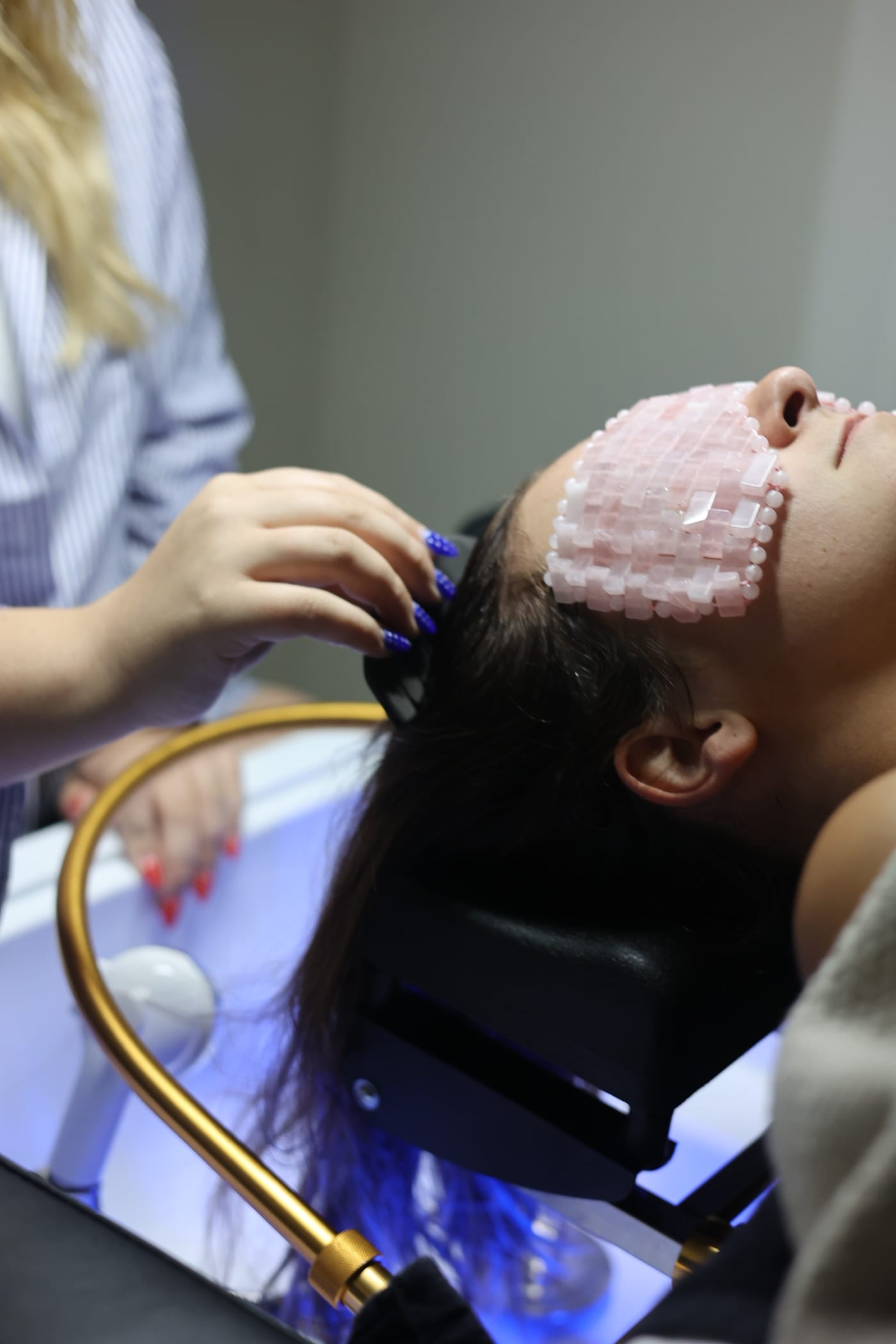 Guest reclined at the gold basin under blue light, wearing a rose-quartz beaded eye mask during the head spa