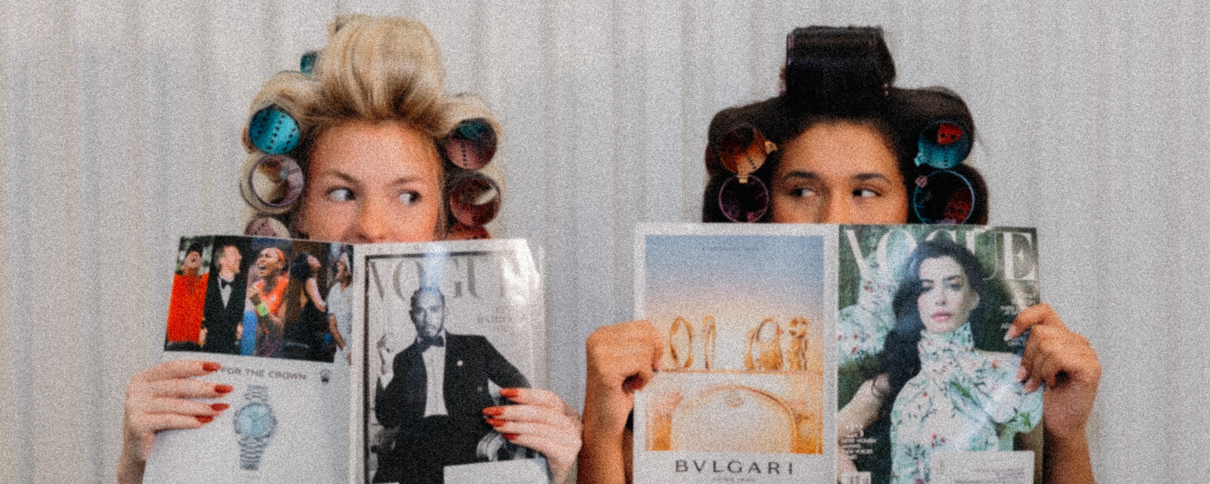 Two guests in colorful hair rollers holding Vogue and Bulgari magazines up over their faces at LŪK House of Beauty