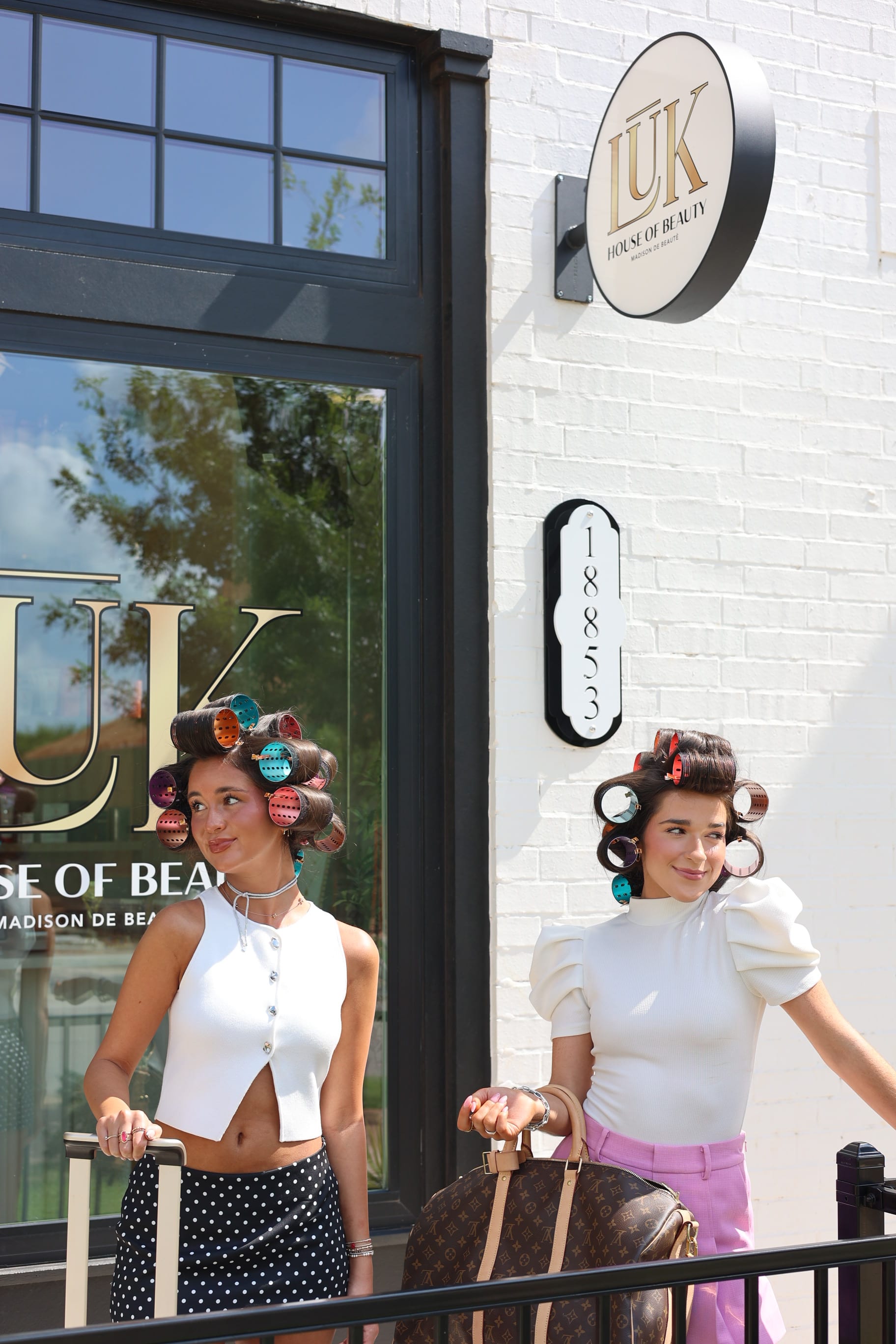 Two women in hair rollers pulling Louis Vuitton luggage outside the LŪK House of Beauty storefront, 18853 Selah Way