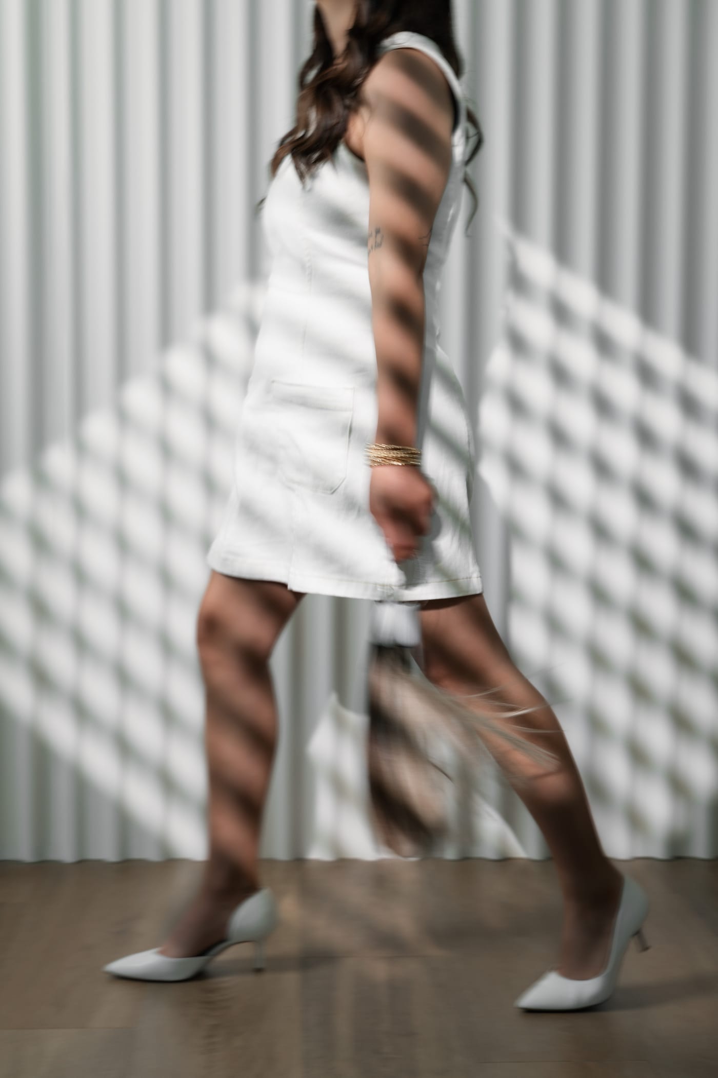 Guest in a white dress and heels walking through bars of window light at LŪK House of Beauty