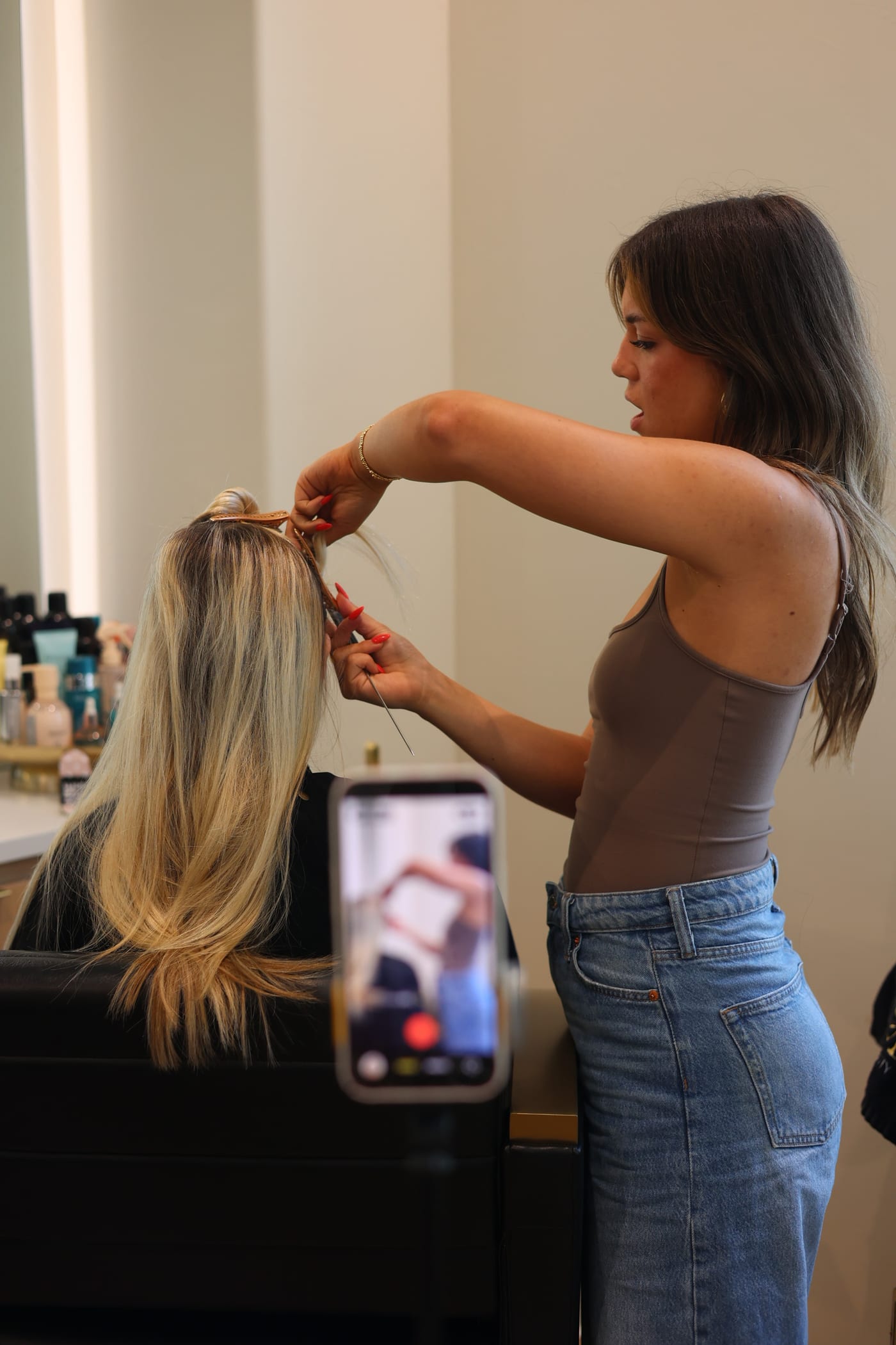 A stylist's hands sectioning a blonde extension weft while an iPhone on a tripod films the work